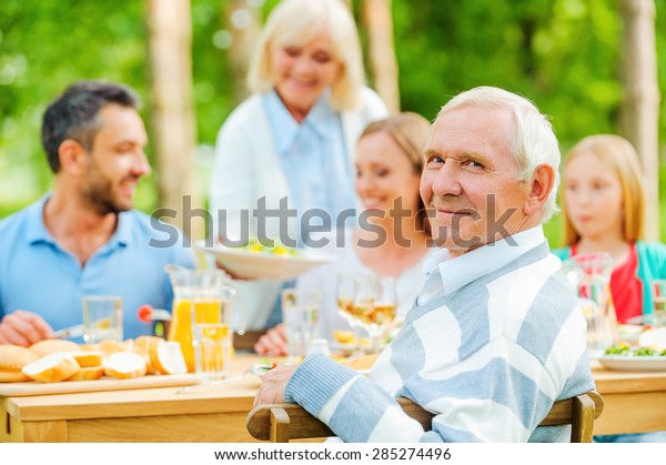Enjoying time with family. Happy family of five people\
sitting at the dining table outdoors while senior man looking over\
shoulder and smiling 