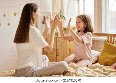 Enjoying time with family. Two caucasian girls, younger and older happy sisters in pajamas playing together while sitting on bed in the morning, young mom and cute little daughter having fun at home