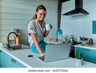 Enjoying time at home. Beautiful young smiling woman working on laptop and drinking coffee while sitting on the countertop in the kitchen at home.