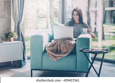 Enjoying time at home. Beautiful young smiling woman working on laptop and drinking coffee while sitting in a big comfortable chair at home 