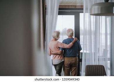 Enjoying time together. Back side portrait of senior woman and man looking on window of balcony while staying embracing at home interior - Powered by Shutterstock