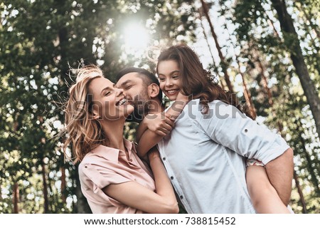 Enjoying time together. Young loving father carrying his smiling daughter and kissing his wife while spending free time outdoors