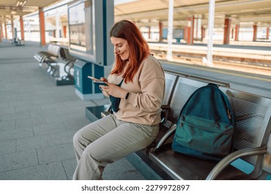 Enjoying travel. Young pretty woman waiting on the station platform with backpack on background electric train using smart phone. Tourist text message and plan route of railway, railroad concept. High
