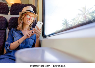 Enjoying travel. Young pretty woman traveling by the train sitting near the window using smartphone.