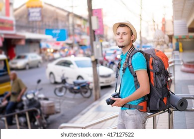 Enjoying Travel. Young Smiling Man With Backpack Holding Camera On Asian Street.