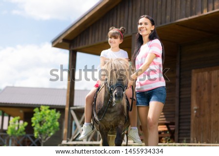 Enjoying weather. Cheerful mother and her daring daughter sitting on the horse enjoying sunny warm weather