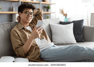 Enjoying Weekend. Handsome Asian Man In Glasses Using Smartphone While Sitting Leaning On Couch At Home, Happy Guy Relaxing In Living Room, Browsing Internet Or Messaging With Friends, Selective Focus