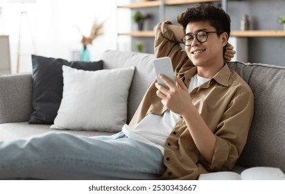 Enjoying Weekend. Handsome Asian Man In Glasses Using Smartphone While Sitting Leaning On Couch At Home, Happy Guy Relaxing In Living Room, Browsing Internet Or Messaging With Friends, Selective Focus - Powered by Shutterstock