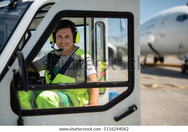 Enjoying work.\
Cheerful man in headphones with microphone closing door of vehicle.\
Airplane on blurred\
background