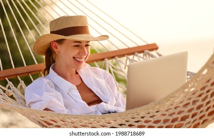 Enjoying Work During Summer Vacation. Young Happy Woman, Successful Female Freelancer In Straw Hat Using Laptop And Smilingwhile Relaxing In The Hammock On The Tropical Beach. Distance Job