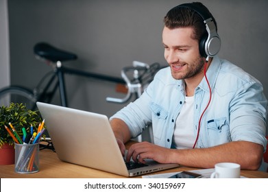 Enjoying Work And Good Music. Cheerful Young Man In Headphones Listening To The Music While Sitting At His Working Place With Bicycle Standing In The Background