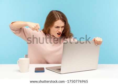 Enraged furious woman in casual clothes roaring madly and punching laptop screen, threatening with fist while talking on video call, online conference at home office. indoor studio shot, isolated