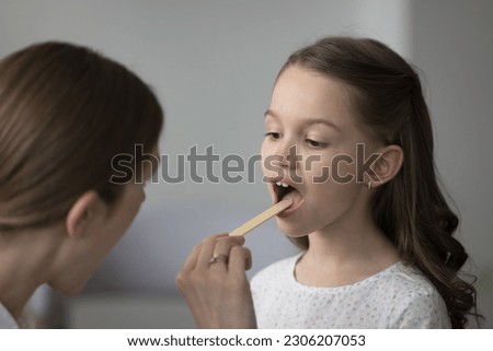 ENT doctor examining little girl throat during visit in clinic, close up. Childs pediatrician holds wooden spatula tongue depressor reviewing patient inflamed tonsils, provide professional medical aid
