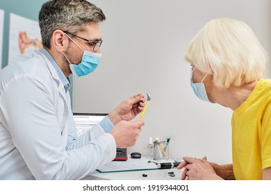 Ent Doctor Wearing A Medical Mask Shows A Hearing Aid To A Senior Woman For Treatment Of Deafness While Coronavirus Epidemic