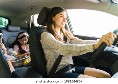 Entertaining My Children. Beautiful Young Mom Smiling While Driving A Car With Her Kids In The Back Watching A Movie On A Tablet With Headphones During Her Errands