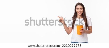 Entertainment, fun and holidays concept. Portrait of cheerful cute young girl waiting for friend in cinema, movie starting soon, pointing finger left inviting come join her, smiling, hold popcorn.