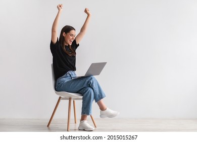 Entertainment And Online Work During Covid. Happy Young Lady Sitting On Chair With Laptop, Raising Her Hands Up, Excited Over Huge Lottery Win Or Business Success In Studio, Copy Space