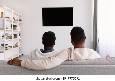 Entertainment. Rear Back View Of Black Young Family Of Two Watching Tv, Sitting On Sofa, Looking At Blank Television Screen On The Wall With Isolated Empty Monitor For Mockup, Banner, Copy Space