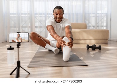 An enthusiastic African American man in athletic wear stretches on a yoga mat for a home workout, recording himself on a phone - Powered by Shutterstock