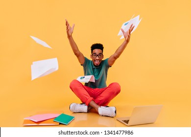 Enthusiastic African Student In Red Pants Fooling Around During Study. Studio Shot Of Smiling Male Freelancer Waving Hands On Yellow Background.