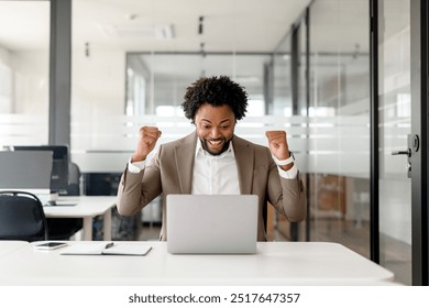 An enthusiastic African-American businessman celebrates a successful moment at his laptop, his fists raised in victory. The joy of achieving business goals and the triumph of entrepreneurial spirit - Powered by Shutterstock