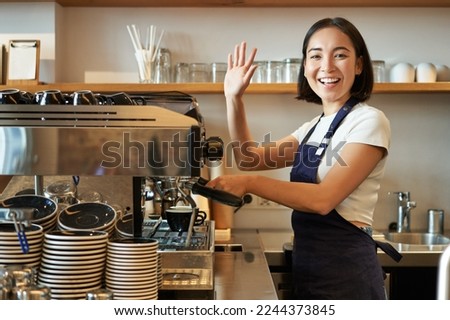 Enthusiastic asian barista, girl using coffee machine, waving at client, saying hello, greeting guests in her cafe.