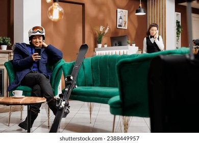 Enthusiastic asian lady using her cell phone and waiting in ski hotel lobby for booking procedure for winter vacation. Traveler in wintersports gear with her smartphone in ski resort lounge area.