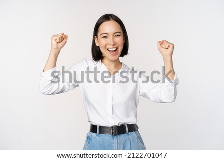 Enthusiastic asian woman rejoicing, say yes, looking happy and celebrating victory, champion dance, fist pump gesture, standing over white background