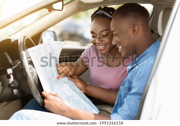 Enthusiastic black couple reading map while\
having car trip together, got lost in the middle of road, side\
view, copy space. Smiling african american young man and woman\
looking for new\
destination