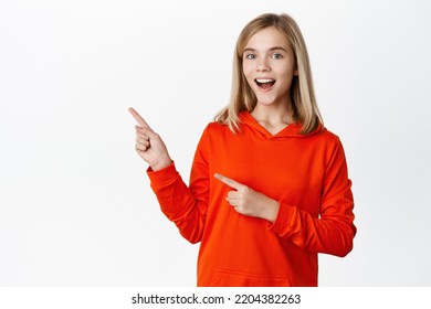 Enthusiastic Blond Little Girl, Teen Kid Pointing Fingers Left And Showing Advertisement, Smiling Happy, Standing Over White Background.