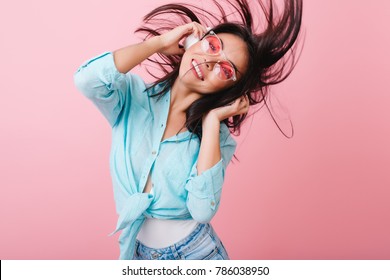 Enthusiastic Brunette Girl Dancing With Hair Waving During Photoshoot In Studio With Pink Interior. Indoor Photo Of Active Hispanic Woman In Headphones And Sunglasses Having Fun.