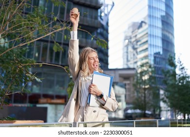 Enthusiastic businesswoman feels upbeat after morning cup of coffee, raise her cup up and dances outside. - Shutterstock ID 2230300051
