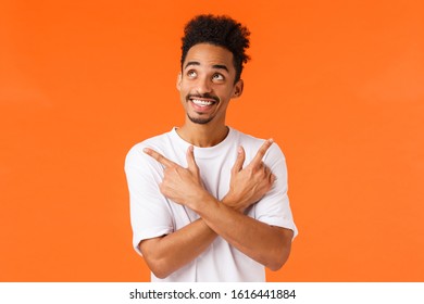Enthusiastic, excited african-american male with moustache, looking up dreamy and carefree, smiling happy, pointing sideways left and right, making choice, decide what buy, orange background