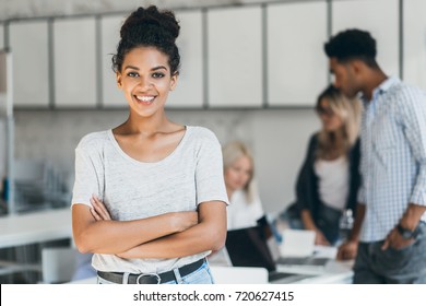 Enthusiastic Female African Student Enjoying Company With Friends In Lecture Hall. Indoor Portrait Of Smiling Black Office Worker Posing With Arms Crossed In Front Of Foreign Colleagues.