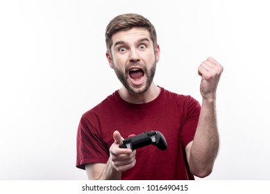 Enthusiastic Gamer. Joyful Bristled Young Man Holding A Video Game Controller And Raising His Hand Triumphantly, Celebrating Victory In Game, While Posing Isolated On A White Background