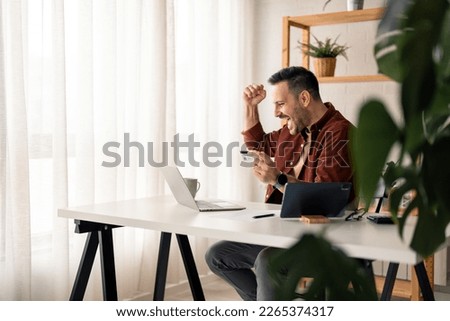 Enthusiastic handsome millennial man feeling excited looking at laptop screen holding credit card, raising fist in yes gesture satisfied with online payment possibilities, rejoicing approved loan.