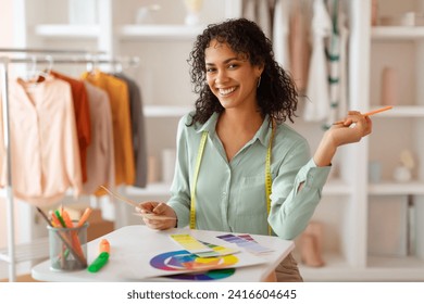 Enthusiastic latin fashion designer with curly hair and measuring tape holds pencil and color wheel, contemplating design choices in her bright studio - Powered by Shutterstock