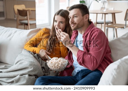 Enthusiastic man and woman eating popcorn watch TV sitting on home sofa enjoying movie. Married relaxed couple wife husband hooked on series. Family pastime, leisure, watch television, resting concept
