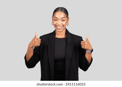 Enthusiastic millennial African American woman in formal attire giving two thumbs up with a radiant smile, exuding positivity and confidence against a neutral grey backdrop, studio - Powered by Shutterstock