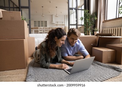 Enthusiastic Millennial Spouses Buyers Renters Of New Flat Lie On Carpet On Floor Among Boxes Packages At Moving Day Use Laptop. Inspired Couple In Love Choose Domestic Interior Design On Pc Online