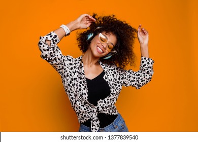 Enthusiastic Mix Race Woman Listening Favorite Music By Earphones And Dancing Over Orange Background. Indoor Black Girl With Close Eyes  Posing In Studio.