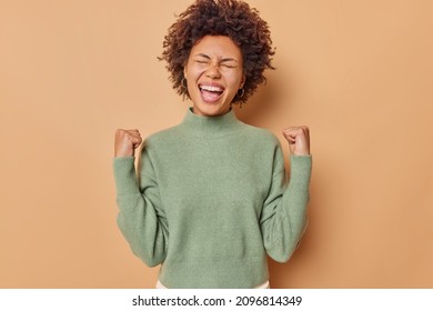 Enthusiastic positive woman with curly hair raises hands up cheers and roots clenches fists as wins something wears casual jumper isolated over beige background achieves goal exclaims loudly - Shutterstock ID 2096814349