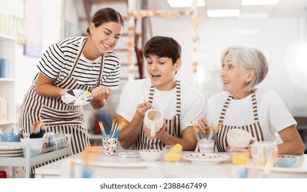 Enthusiastic senior female teacher sharing experience pottery craftsmanship, assisting happy young guy and girl, painting ceramic mugs and plates at table in workshop - Powered by Shutterstock