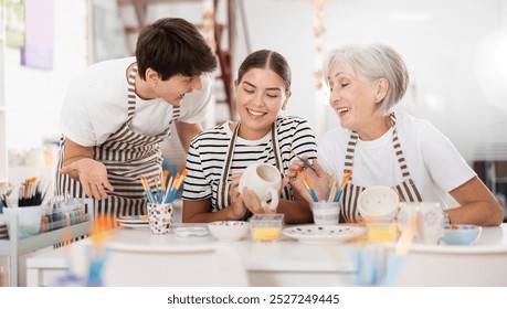 Enthusiastic senior female teacher sharing experience pottery craftsmanship, assisting happy young guy and girl, painting ceramic mugs and plates at table in workshop - Powered by Shutterstock