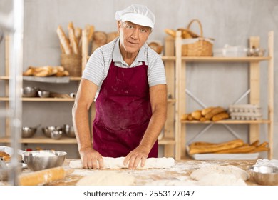 Enthusiastic skilled aged baker wearing maroon apron and white hairnet working in cozy artisanal bakery, rolling out soft dough, shaping baguette on floured wooden table, preparing for baking - Powered by Shutterstock