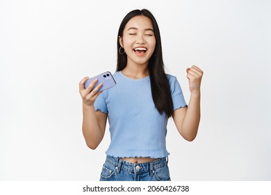 Enthusiastic Smiling Asian Woman Winning On Mobile Phone Video Game, Looking Satisfied And Relieved, Standing In T-shirt Over White Background