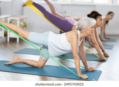 Enthusiastic sporty senior lady performing set of exercises with resistance band during group pilates class in light airy fitness studio - Powered by Shutterstock