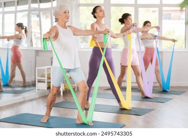 Enthusiastic sporty senior lady performing set of exercises with resistance band during group pilates class in light airy fitness studio - Powered by Shutterstock