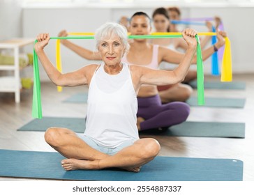 Enthusiastic sporty senior lady performing set of exercises with resistance band during group pilates class in light airy fitness studio - Powered by Shutterstock