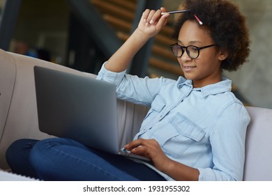 Enthusiastic Teen Schoolgirl In Glasses With Pencils In Her Hair Looking Focused While Using Laptop For Studying, Sitting On The Couch At Home